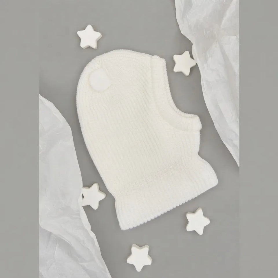 A white ribbed knit baby bonnet or hat laid flat on a gray background, surrounded by white star-shaped decorative pieces and crumpled white paper.