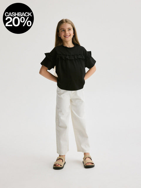 Young girl wearing a black top with ruffles and white pants on a light gray background