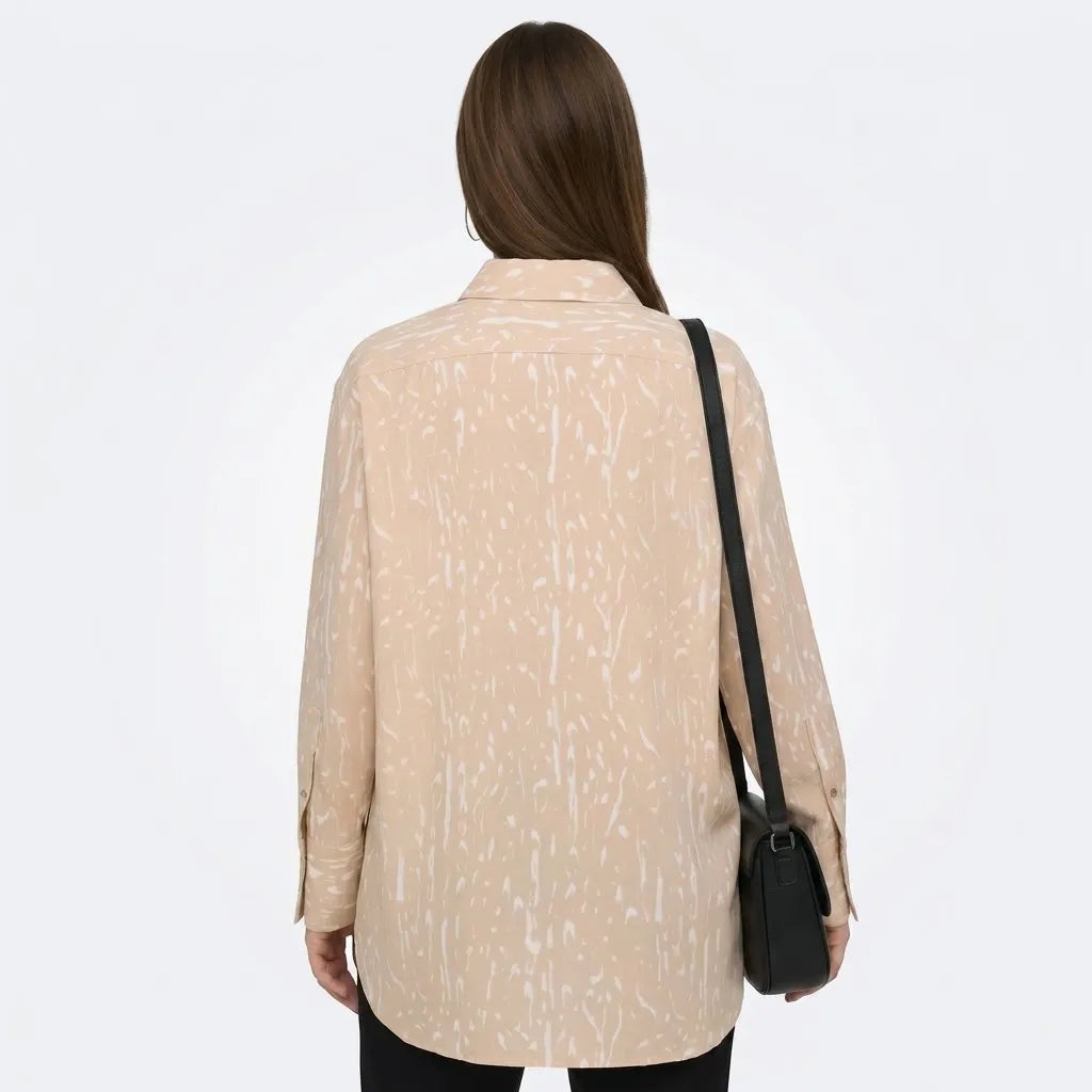 A woman viewed from behind wearing a beige long-sleeve button-up shirt with a white abstract brushstroke pattern, paired with black pants and carrying a black shoulder bag against a plain white background.
