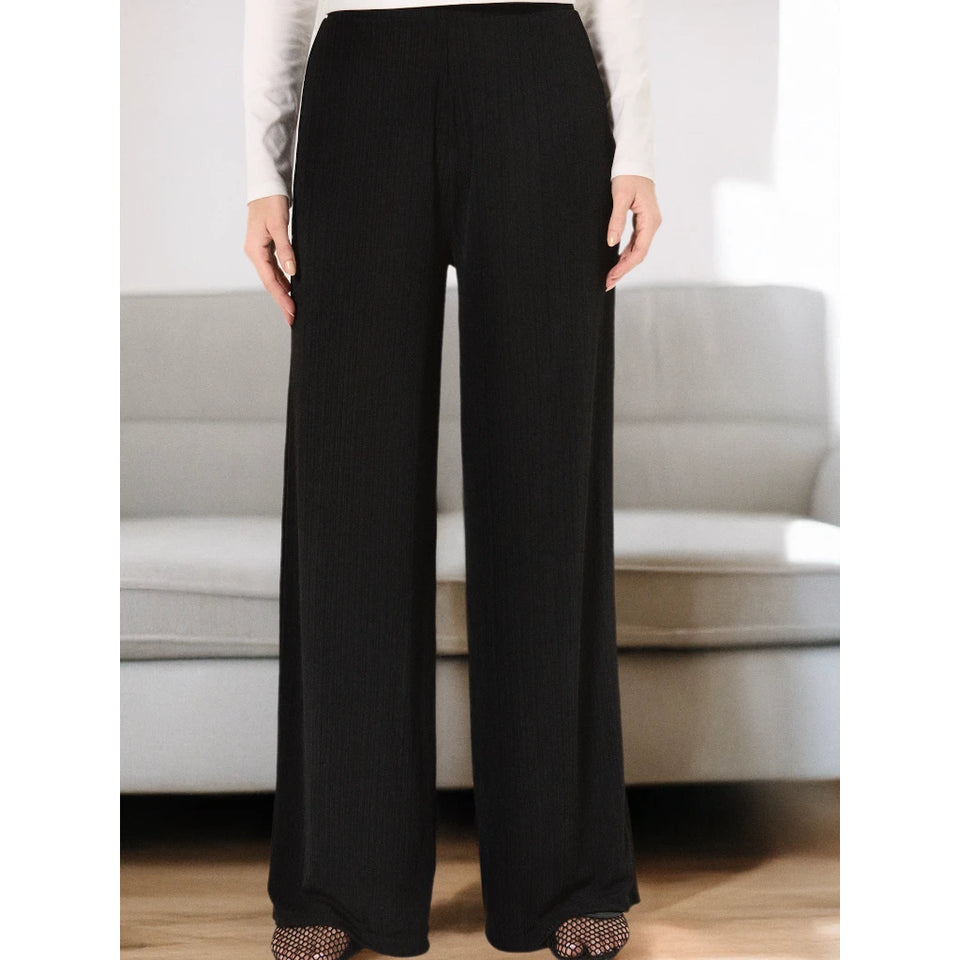 Black wide-leg pleated trousers shown on a model in a living room setting with a light gray sofa in the background.