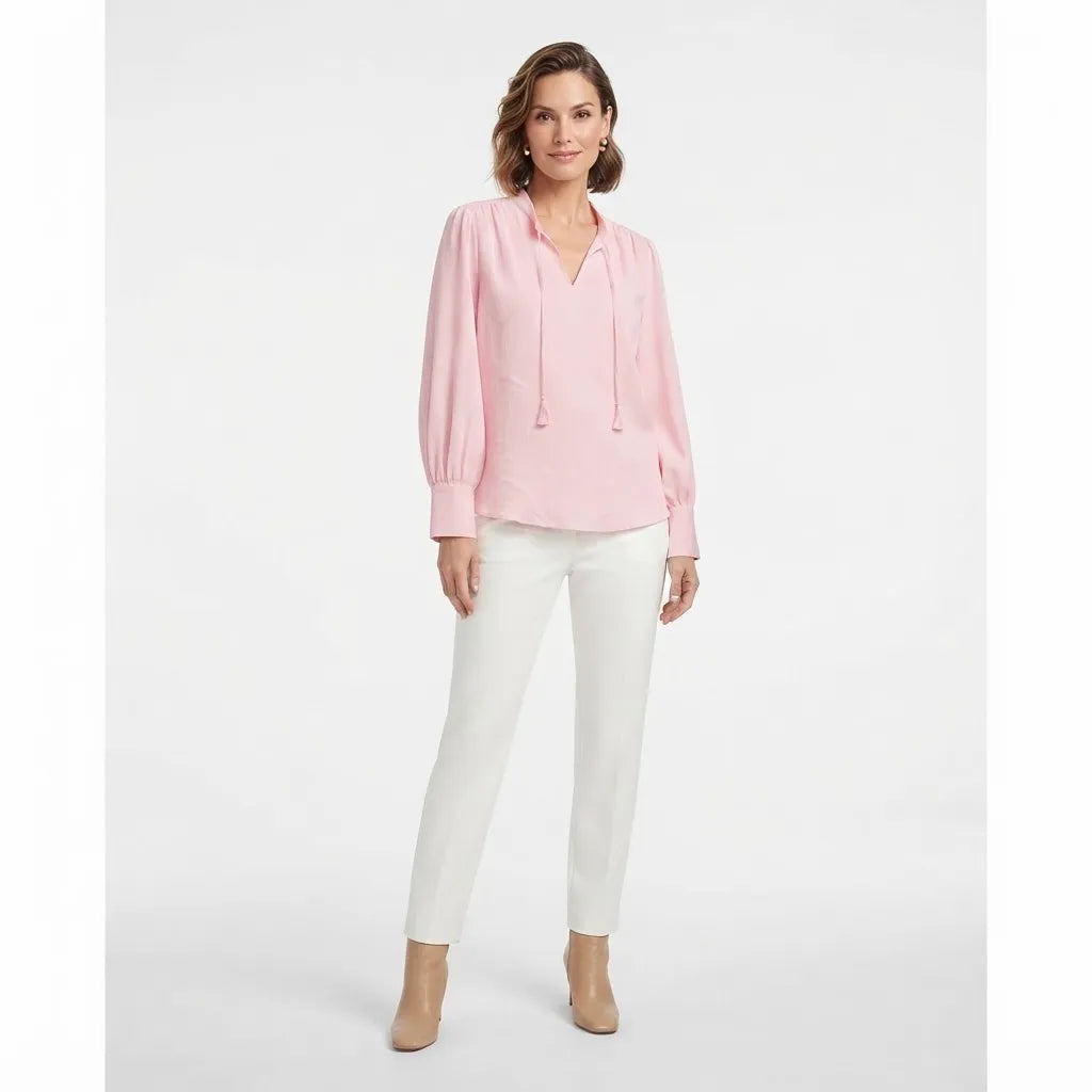 A woman modeling a light pink long-sleeve blouse with a split neckline and drawstring ties, paired with white pants and beige heels, against a plain light gray studio background.