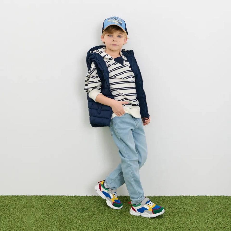 A young boy modeling a navy blue puffer vest over a navy and white striped long-sleeve shirt, paired with light blue jeans, a blue cap, and colorful sneakers, standing against a white wall with artificial green turf at the bottom.