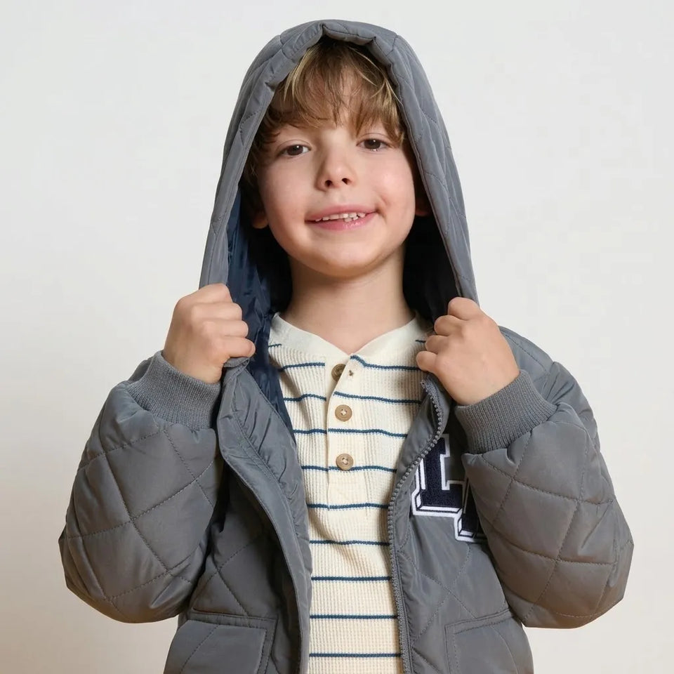 A young boy wearing a gray quilted puffer jacket with a hood over a cream and navy striped henley shirt, posing against a light off-white background.