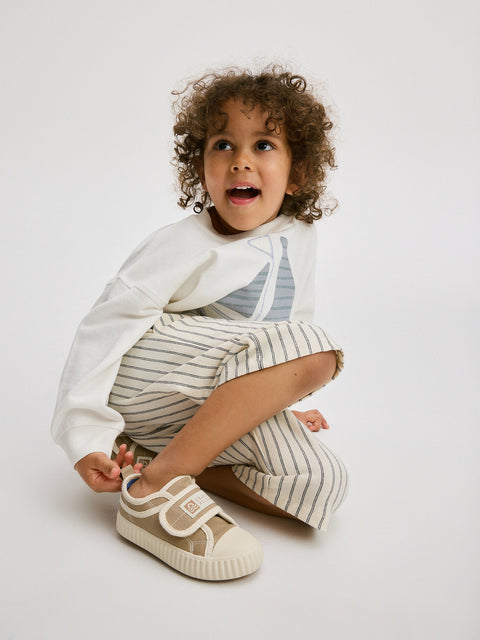 Child wearing a white long-sleeve shirt with a sailboat design and striped pants, sitting on a light gray background.