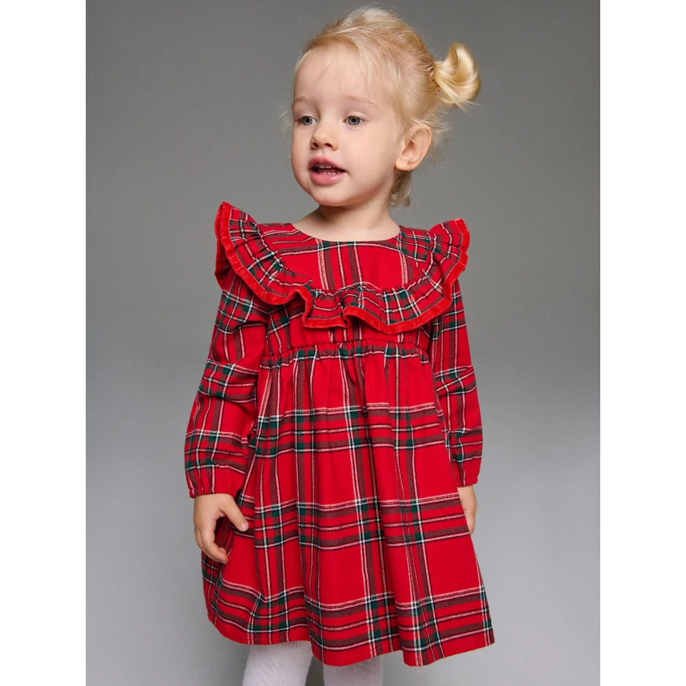 A young toddler girl wearing a red long-sleeve dress with a green and black plaid pattern, ruffled collar, and matching bow, standing against a solid gray studio background.