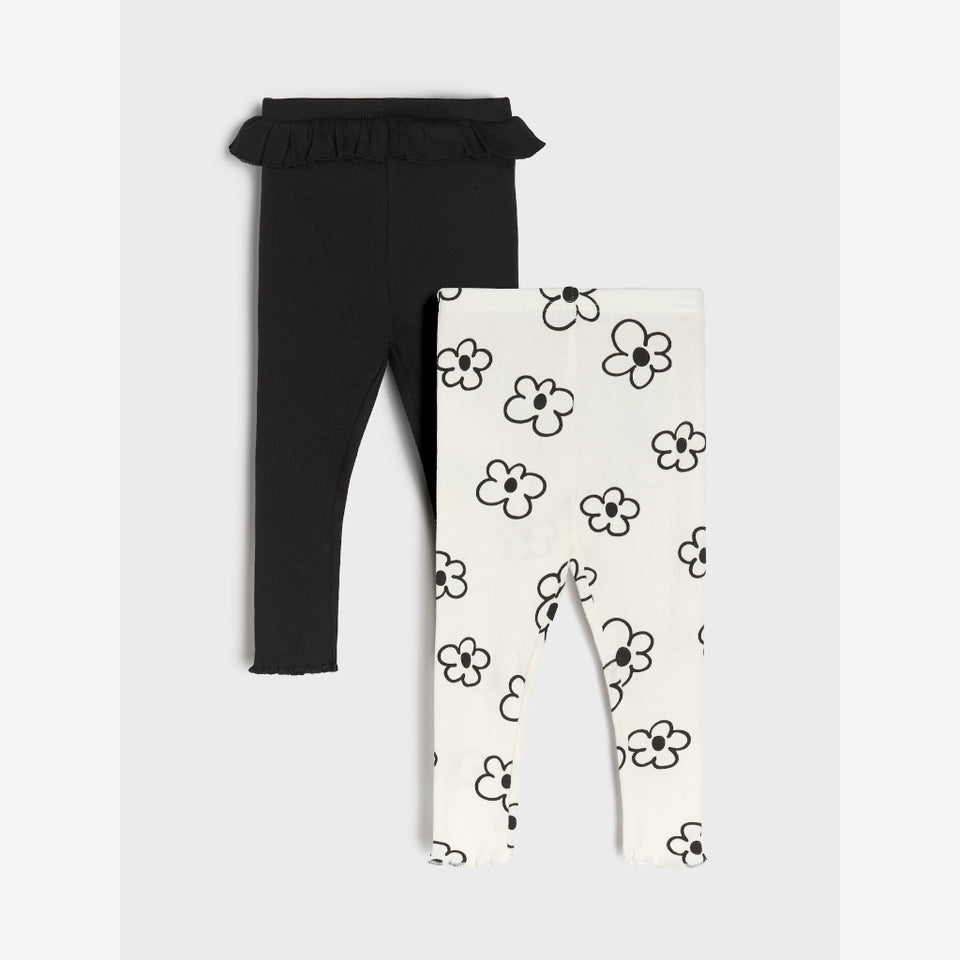 Two pairs of toddler leggings laid flat on a light gray background: one pair is solid black with a ruffled waistband, and the other pair is off-white with a black daisy floral print.