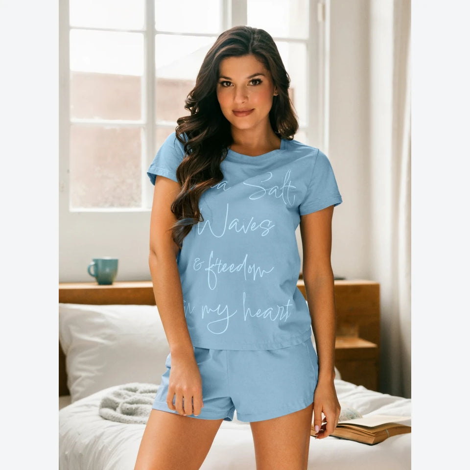 A woman modeling a light blue two-piece pajama set with a matching graphic t-shirt and shorts in a bright bedroom setting.