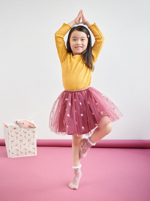 Young girl in a yellow sweater and pink skirt posing on a pink floor with a white background