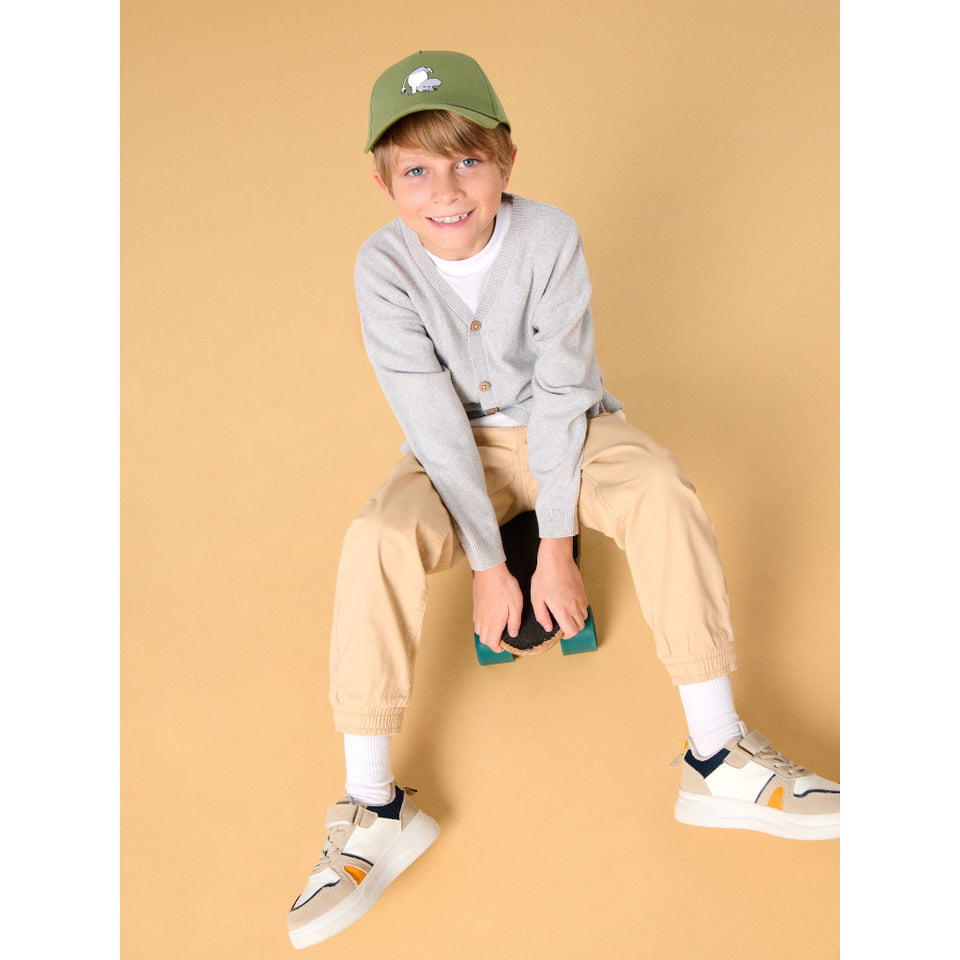 A young boy wearing a gray cardigan, beige trousers, white sneakers, a white t-shirt, and an olive green cap sits on a small stool against a solid beige background.
