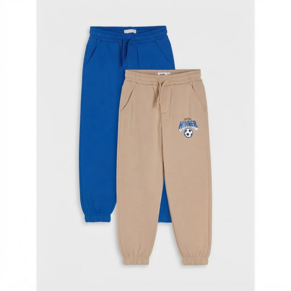 Two pairs of kids' jogger sweatpants, one in royal blue and one in beige, displayed against a light gray background. The beige pair features a small embroidered soccer-themed logo on the left thigh.