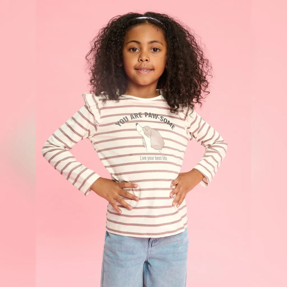 A young girl with curly black hair stands against a solid pink background wearing a long-sleeve white shirt with thin brown horizontal stripes and a graphic print that reads 'YOU ARE PAW-SOME' with a paw illustration. She is also wearing light blue jeans.