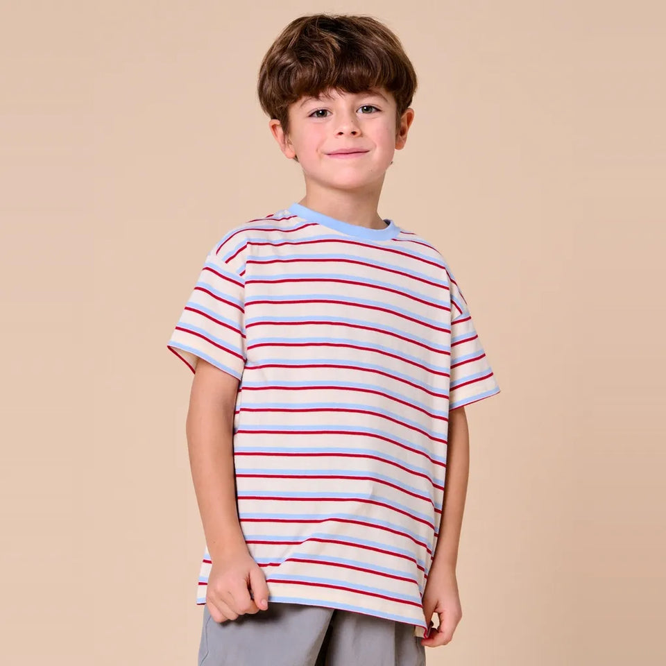 A young boy with brown hair wearing a white short-sleeve t-shirt with thin red and blue horizontal stripes, paired with gray pants, standing against a solid beige background.