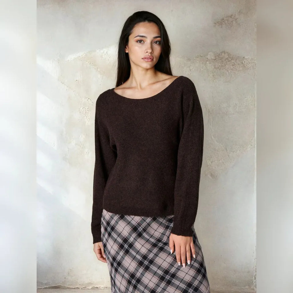 A woman modeling a dark brown knit sweater with a wide boat neckline and long sleeves, paired with a plaid skirt, standing against a textured concrete wall.