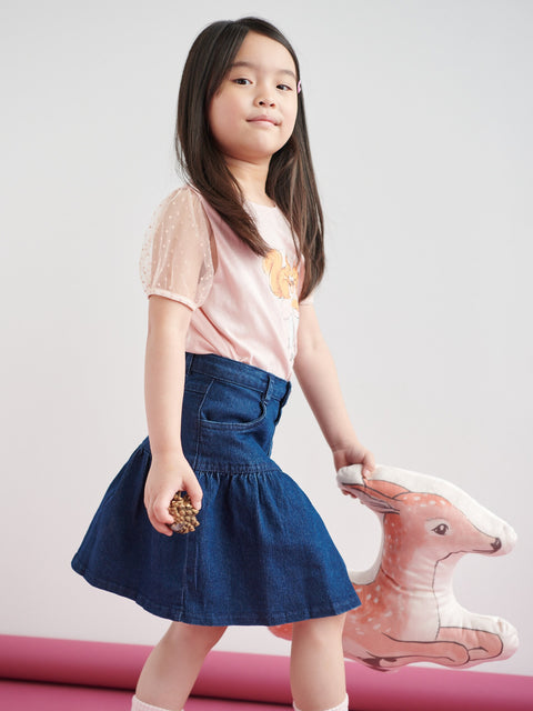 Young girl in a pink shirt and blue skirt holding a toy deer against a light gray background