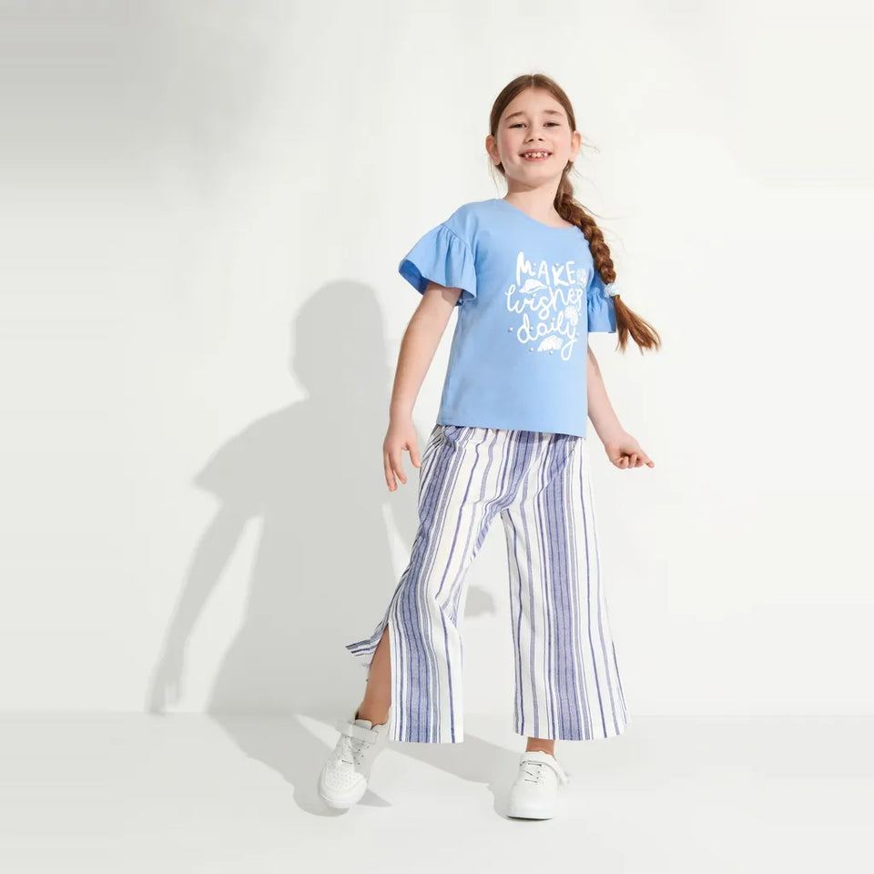 A young girl with a long braided ponytail stands in a studio wearing a light blue graphic t-shirt and blue-and-white vertically striped wide-leg pants with side slits, paired with white sneakers.