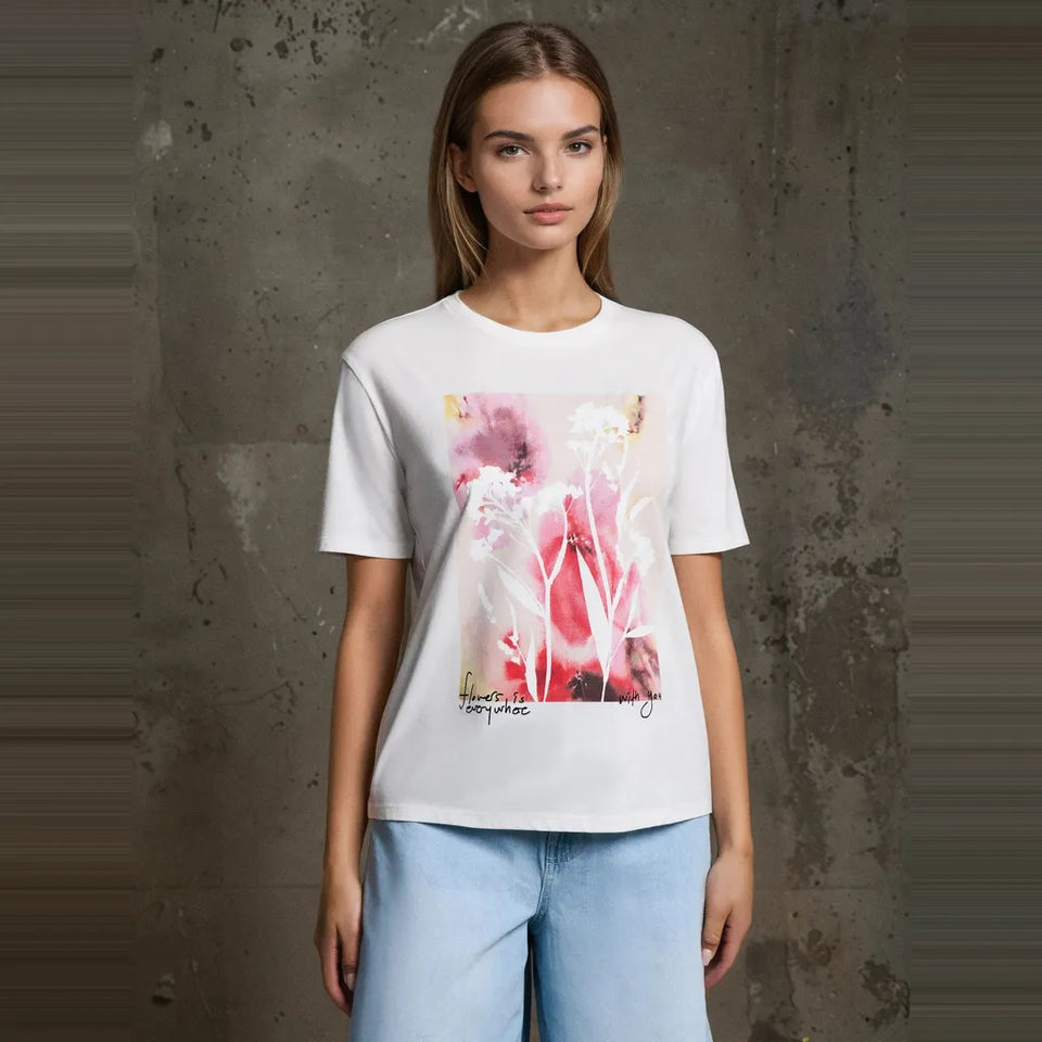 A woman modeling a white short-sleeve t-shirt with a pink and red abstract floral graphic print on the front, paired with light blue jeans, set against a textured gray concrete wall.