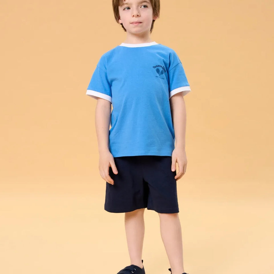 A young boy modeling a light blue ringer t-shirt with white trim and a dark navy shorts, standing against a solid beige background.