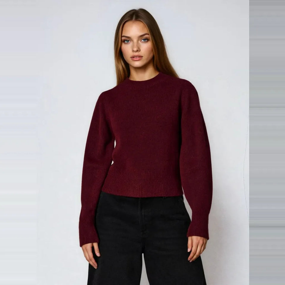 A woman modeling a dark burgundy knit crewneck sweater with black pants, standing against a light gray studio background.
