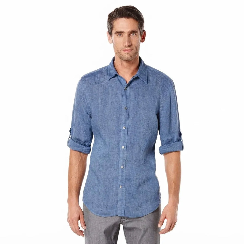 A man wearing a blue chambray long-sleeve button-down shirt with rolled-up sleeves and gray trousers, standing against a plain white background.