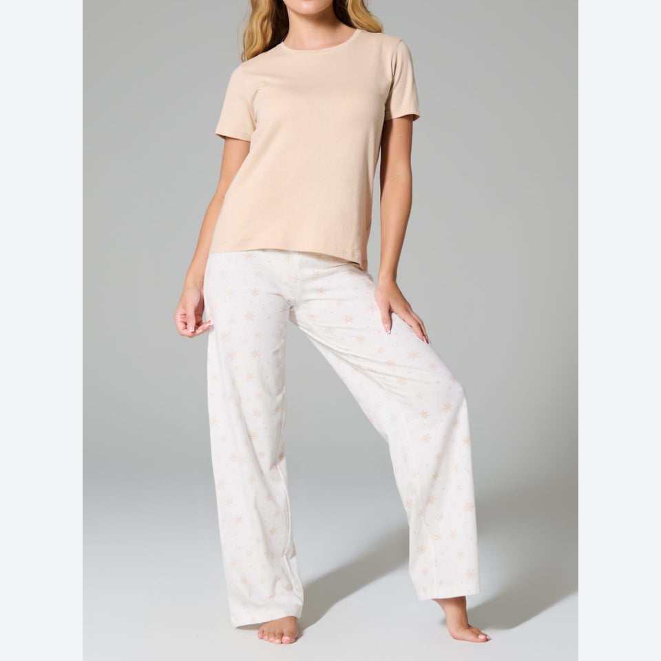 A woman modeling a two-piece pajama set consisting of a short-sleeve crewneck t-shirt in light beige and wide-leg pants in white with a subtle beige pattern, set against a plain gray studio background.
