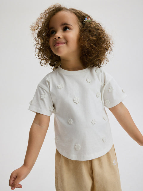 Child wearing a white shirt with floral patterns and beige pants on a plain background