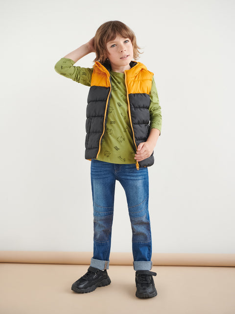 Child wearing a green shirt, blue jeans, and a multicolored puffer vest on a beige background