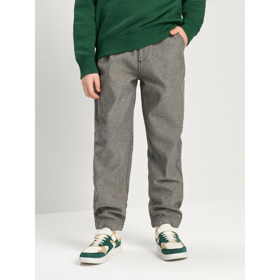 A child wearing gray heathered jogger pants, a green sweatshirt, and green and white sneakers, standing against a light gray studio background.