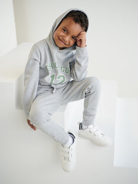 Child wearing a gray hoodie and pants with 'CHIC CO' text, sitting on a white surface.