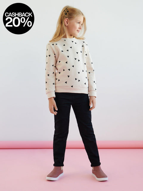Young girl wearing a light pink sweater with black heart patterns and dark pants on a white background.