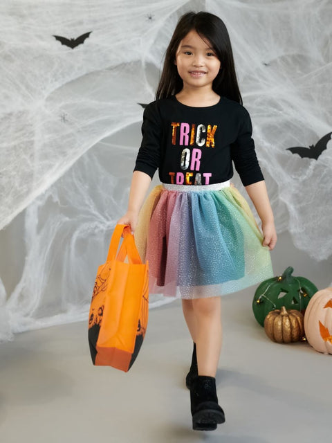Child in Halloween-themed outfit with colorful skirt and 'Trick or Treat' shirt, holding an orange bag against a spider web background.