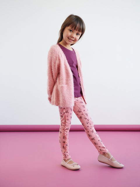 Young girl wearing a pink cardigan, purple shirt, and floral leggings on a pink floor with a white background