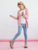 Young girl wearing a pink cardigan, white shirt with a cat design, blue jeans, and brown boots on a pink floor with a white background.