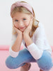 Young girl wearing a pink headband and white shirt with blue jeans, sitting on a pink surface.
