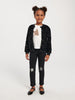 Young girl wearing a black sequin jacket, white shirt with star design, and black pants with star patches on a white background.