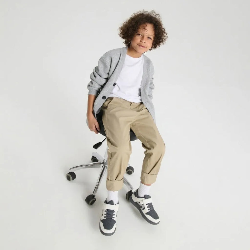 A child with curly hair sits on a black office chair, wearing a light gray cardigan over a white t-shirt, beige cuffed pants, white socks, and black and white sneakers, against a plain light gray studio background.