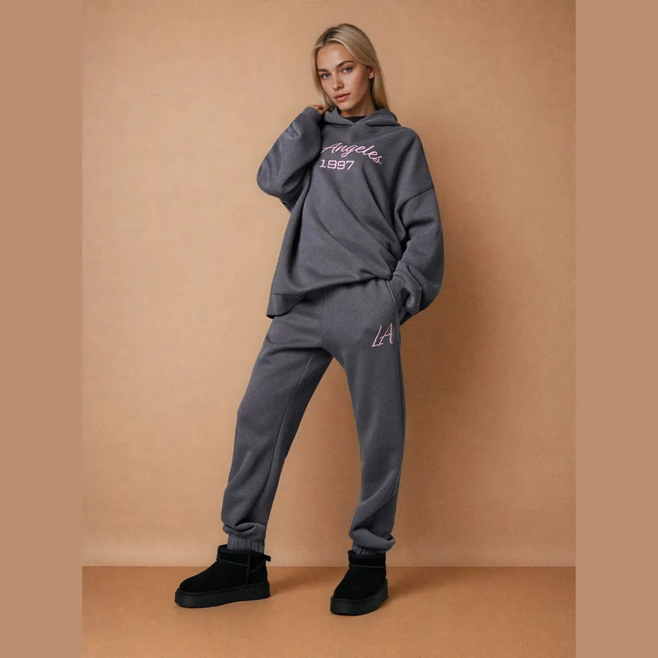 A woman modeling a dark gray lounge set consisting of a sweatshirt and jogger pants with pink embroidered details, standing against a solid tan background.