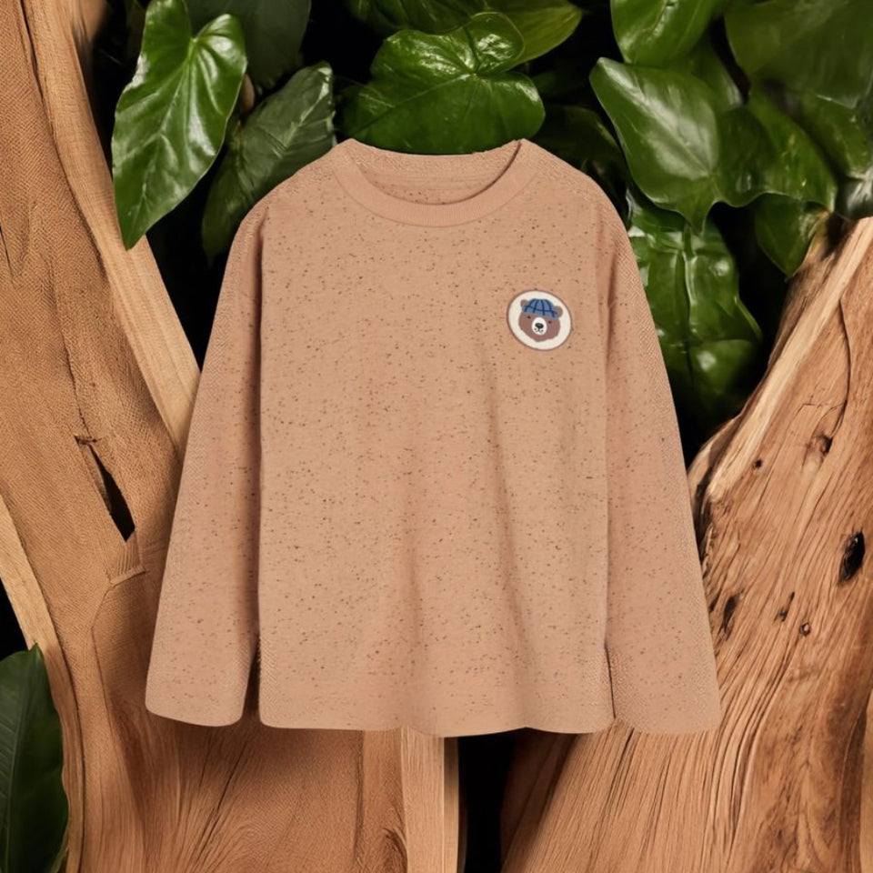 A beige long-sleeve crewneck sweatshirt with a small circular graphic patch on the left chest, displayed on a rustic wood background with green leaves.