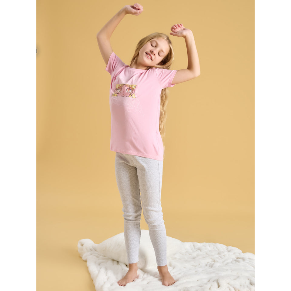 A young girl with long blonde hair is wearing a light pink short-sleeve t-shirt with a small graphic on the chest and light gray heathered leggings. She is standing barefoot on a white textured blanket against a solid warm yellow studio background.