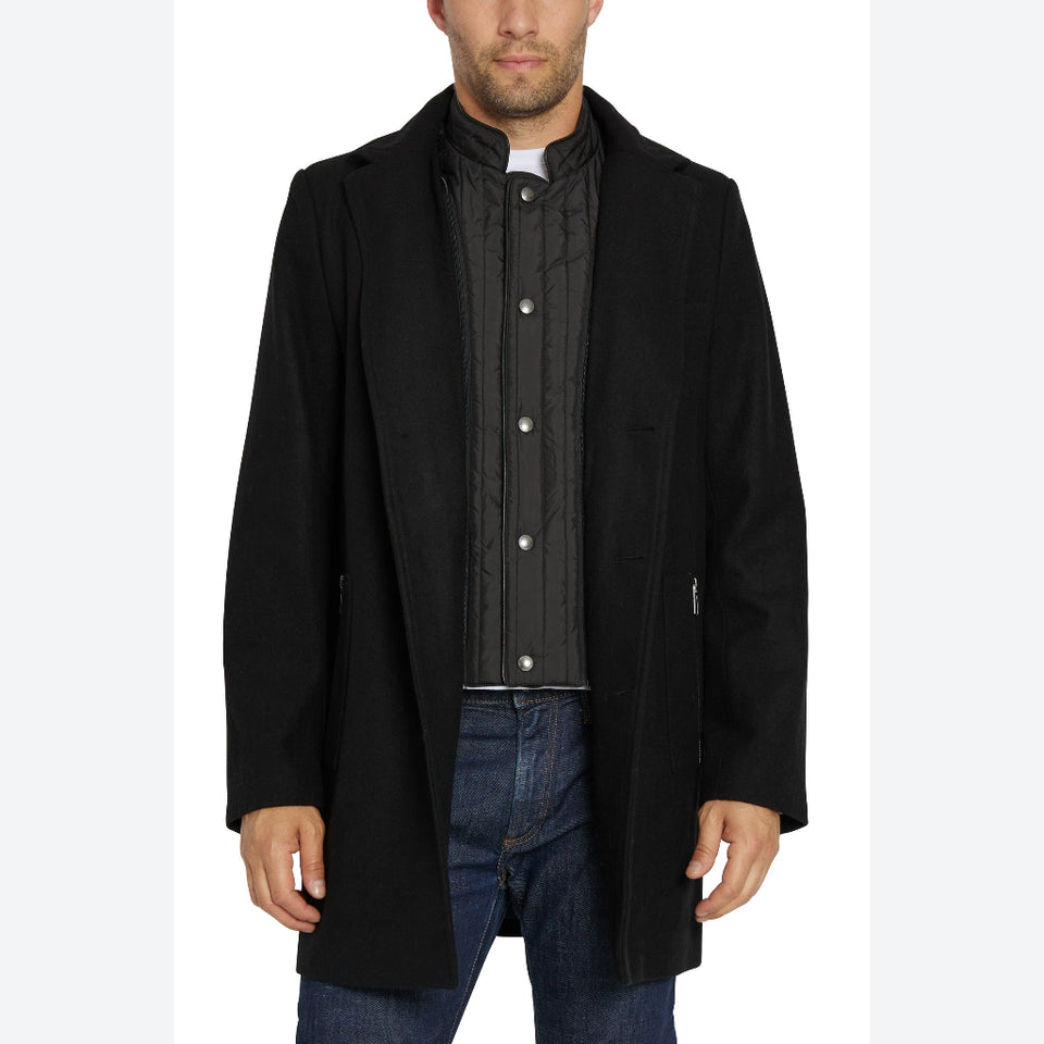 A man wearing a black mid-length overcoat over a black quilted button-front shirt and blue jeans, photographed against a plain white studio background.