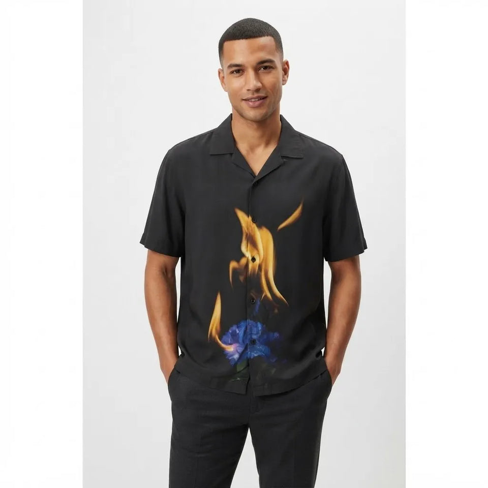 A man wearing a black short-sleeve button-up shirt with a graphic print of a blue flower burning with orange flames, paired with black pants, against a light gray background.