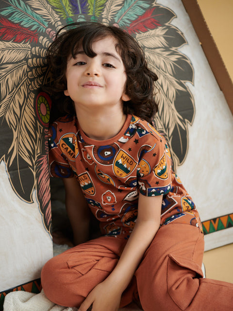 Child wearing a patterned shirt and brown pants sitting against a decorative wall.