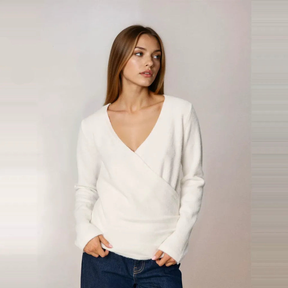 A woman wearing a white long-sleeve wrap-style sweater and dark blue jeans, standing against a light gray studio background. A small 'AI' label appears in the top-left corner.