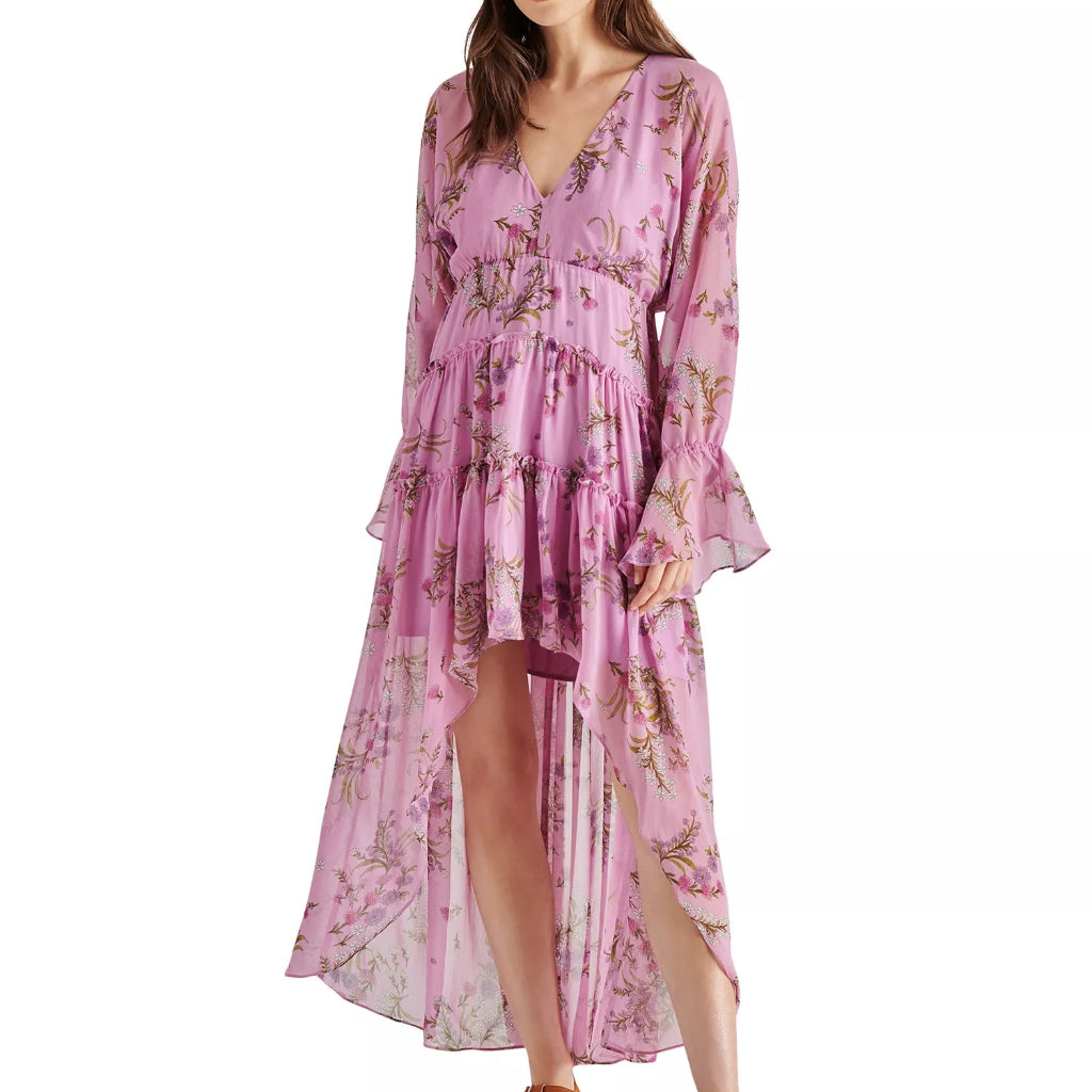 A woman modeling a long-sleeved pink floral midi dress with a tiered ruffle skirt and high-low hemline, paired with brown platform sandals against a plain white background.