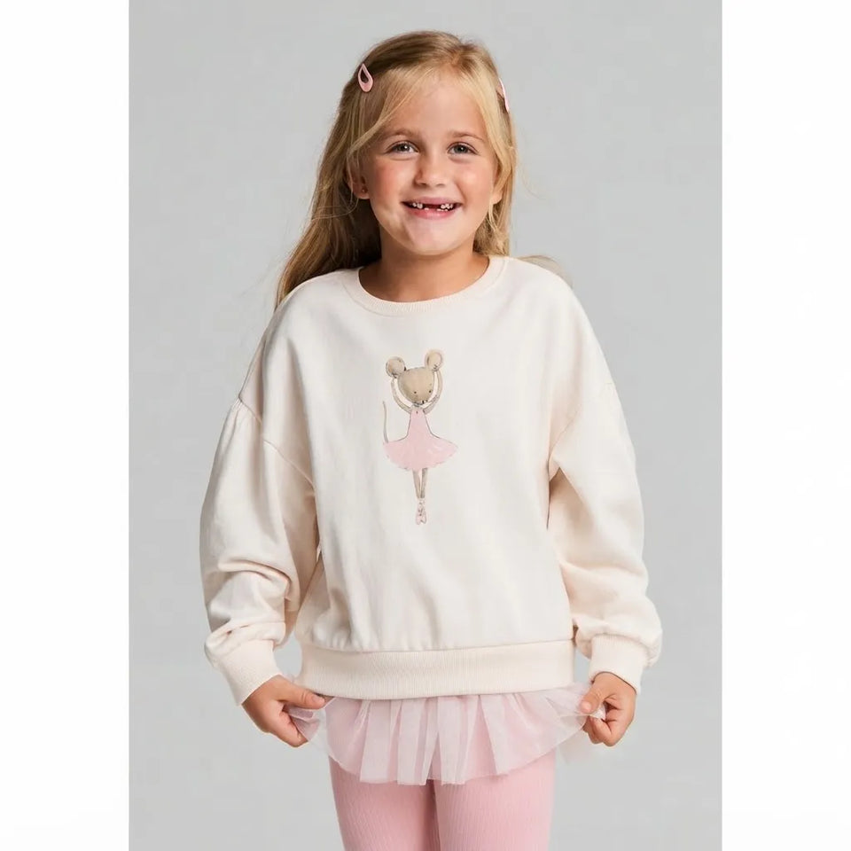 A young girl with blonde hair and pink hair clips is smiling while wearing a cream-colored sweatshirt with a ballerina graphic, a light pink tutu skirt, and pink tights, set against a plain light gray background.