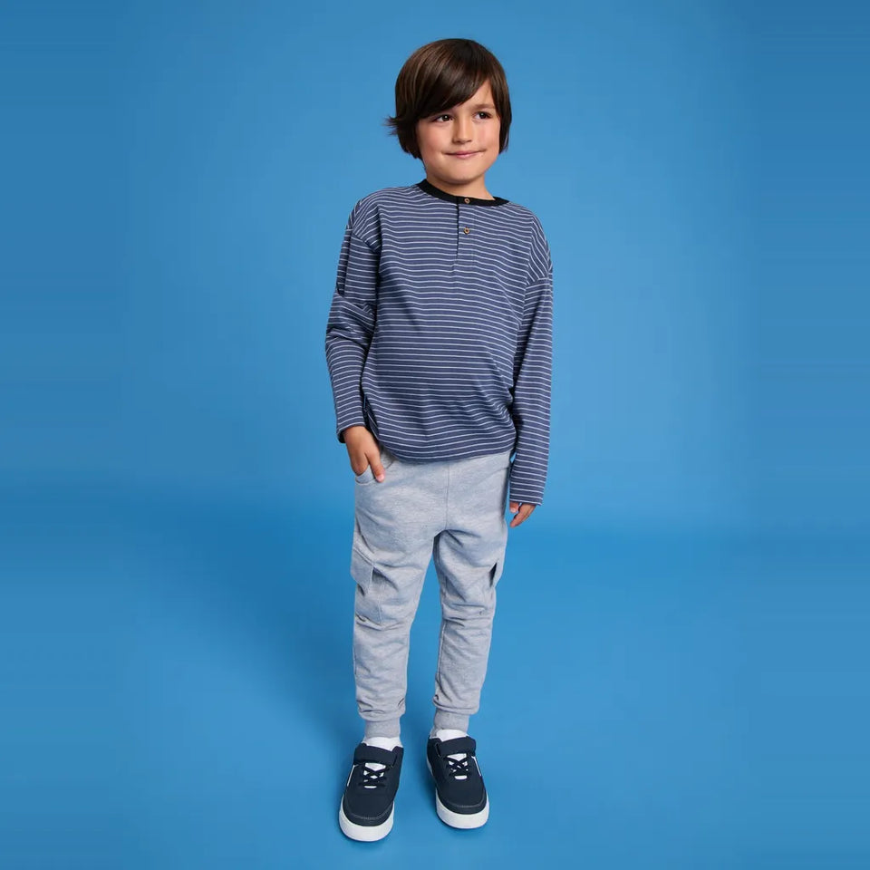 A young boy stands against a solid blue studio background wearing a navy and gray striped long-sleeve shirt, light gray jogger pants, and navy sneakers.