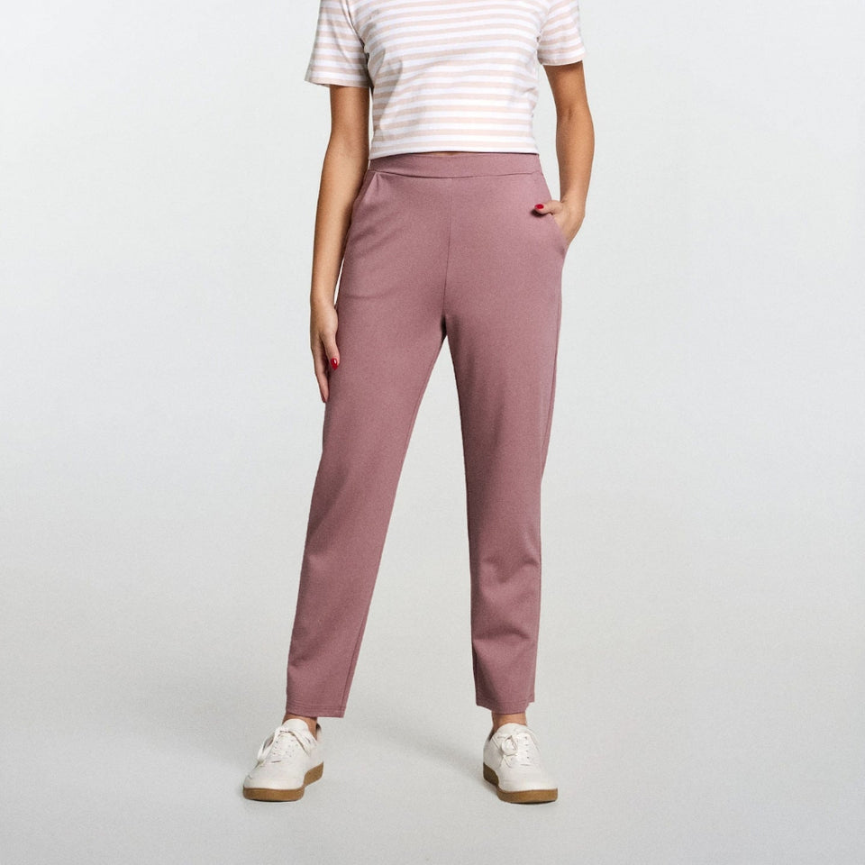A woman modeling a white short-sleeve t-shirt with thin pink horizontal stripes and high-waisted, straight-leg dusty rose pants, paired with white sneakers, against a light gray studio background.