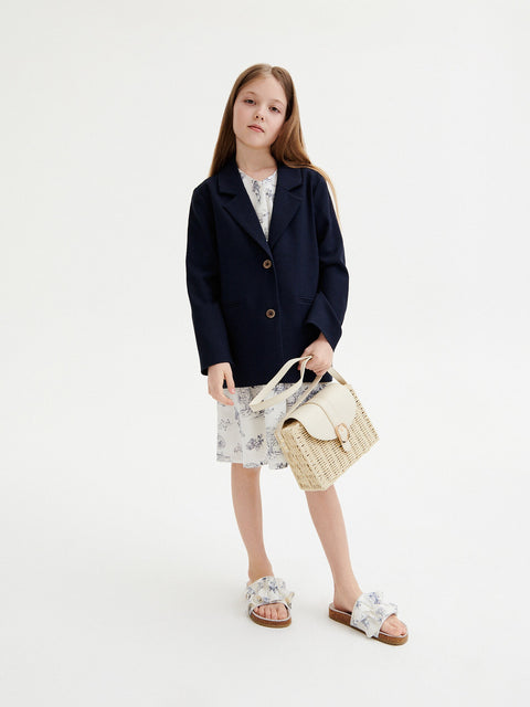 Young girl wearing a navy blazer, floral dress, and holding a straw bag on a white background