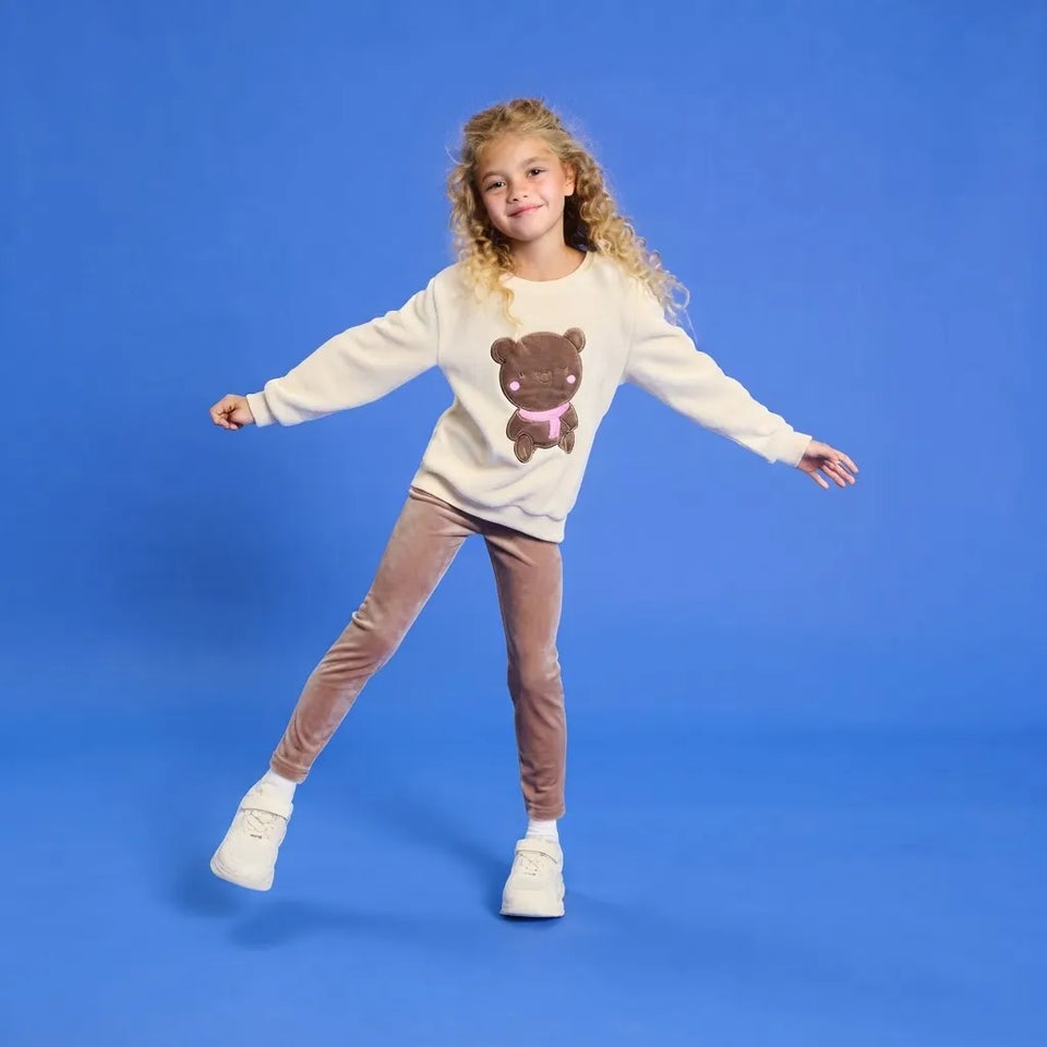 A young girl with curly blonde hair is posing in a studio setting against a solid blue background. She is wearing a cream-colored sweatshirt with a brown teddy bear graphic, brown pants, and white sneakers.