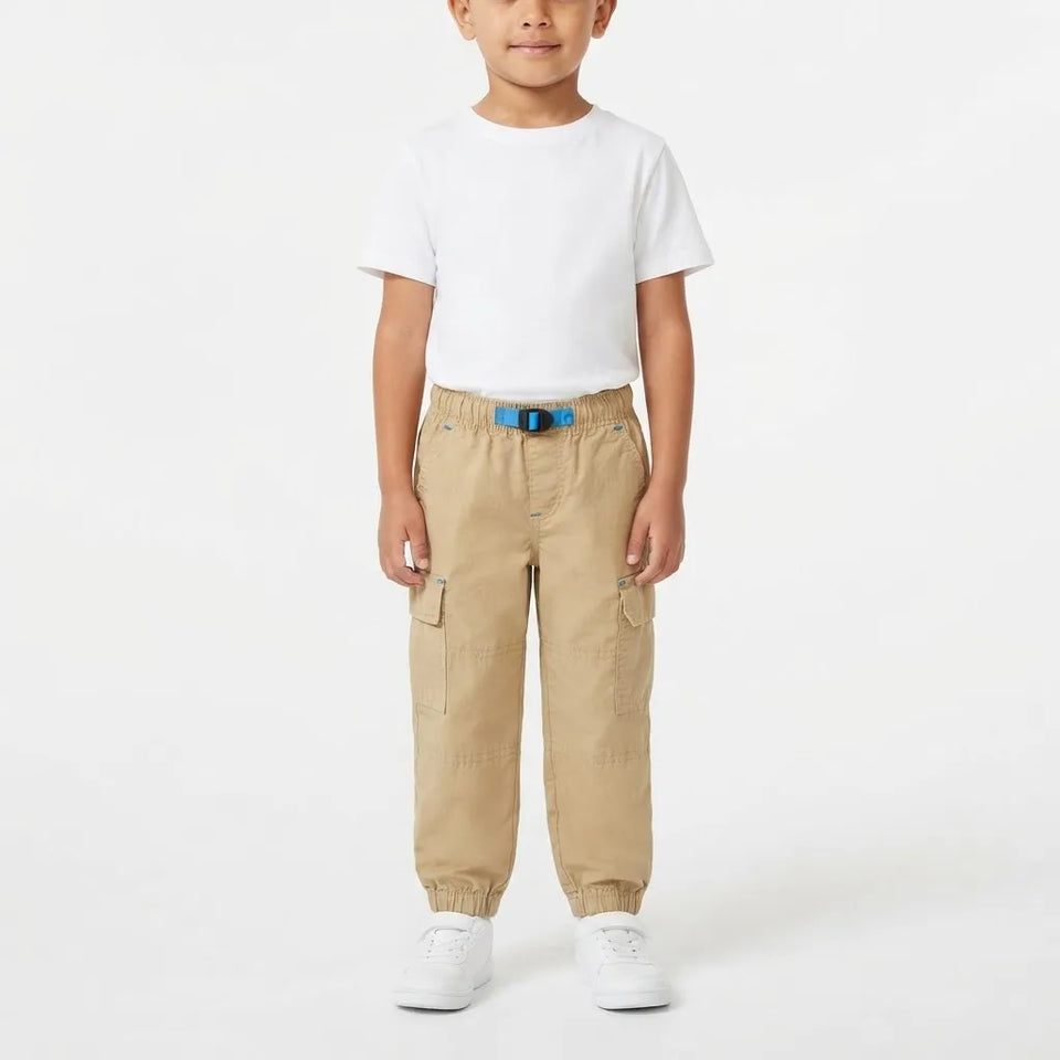 A young child standing against a plain white studio background, wearing a white short-sleeve T-shirt, tan cargo jogger pants with a blue belt, and white sneakers.