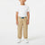 A young child standing against a plain white studio background, wearing a white short-sleeve T-shirt, tan cargo jogger pants with a blue belt, and white sneakers.